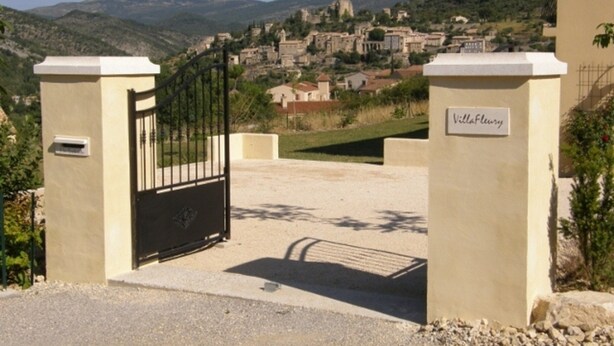 Ein verschlossenes Tor mit dem Schild 'VillaFlurry', das zu einem Dorf mit historischen Gebäuden und Hügeln im Hintergrund führt.