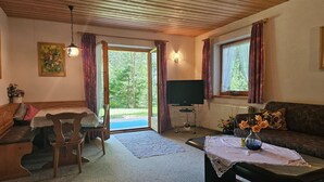 Een gezellige woonkamer met een houten plafond, een eettafel met stoelen, een bank, een televisie en een raam met gordijnen.