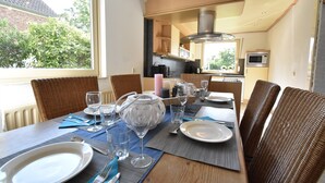 Un comedor con una mesa preparada para cuatro personas, con platos, vasos y cubiertos. Hay sillas de mimbre y se ve la cocina al fondo.