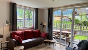 Ein gemütliches Wohnzimmer mit einer Couch, zwei kleinen Tischen und einem Sessel. Große Fenster bieten Blick auf eine Holzterrasse und ein Haus.