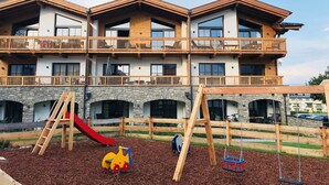 A multi-story building with balconies, a playground with a slide and swings, and a stone facade. - Cozy Apartment in Piesendorf (Piesendorf)