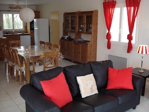 A living room with a black sofa, red cushions, a dining table with chairs, a kitchen area with wooden cabinets, and a red curtain. - Panoramic Poolside Villa (La Croix-Blanche)