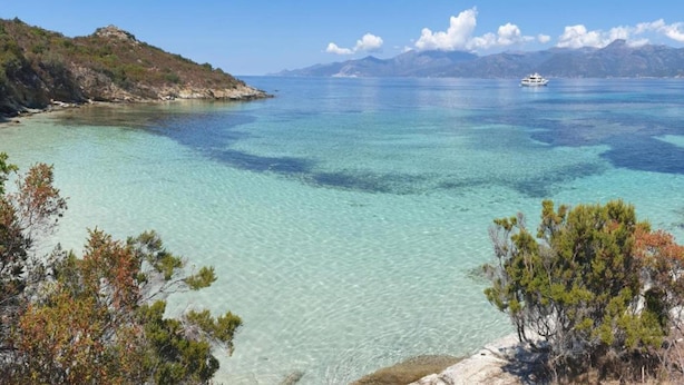 A coastal landscape with clear turquoise waters, a boat in the distance, and a rocky shoreline with sparse vegetation.