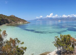 A coastal landscape with clear turquoise waters, a boat in the distance, and a rocky shoreline with sparse vegetation. - Riverside Home in Folelli (Taglio-Isolaccio)