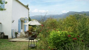 A patio with a table and chairs, a white building with blue shutters, and a view of a green landscape with hills.