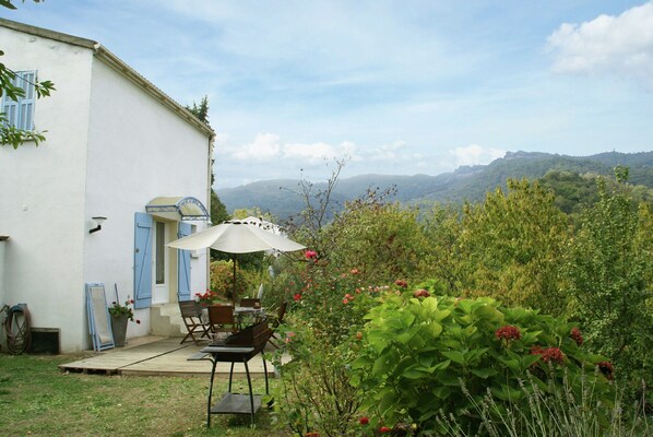 Un patio con una mesa y sillas, un edificio blanco con contraventanas azules, y vistas a un paisaje verde con colinas.