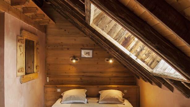 Een houten kamer met een schuine kap, een bed, een stoel en een klein raam.