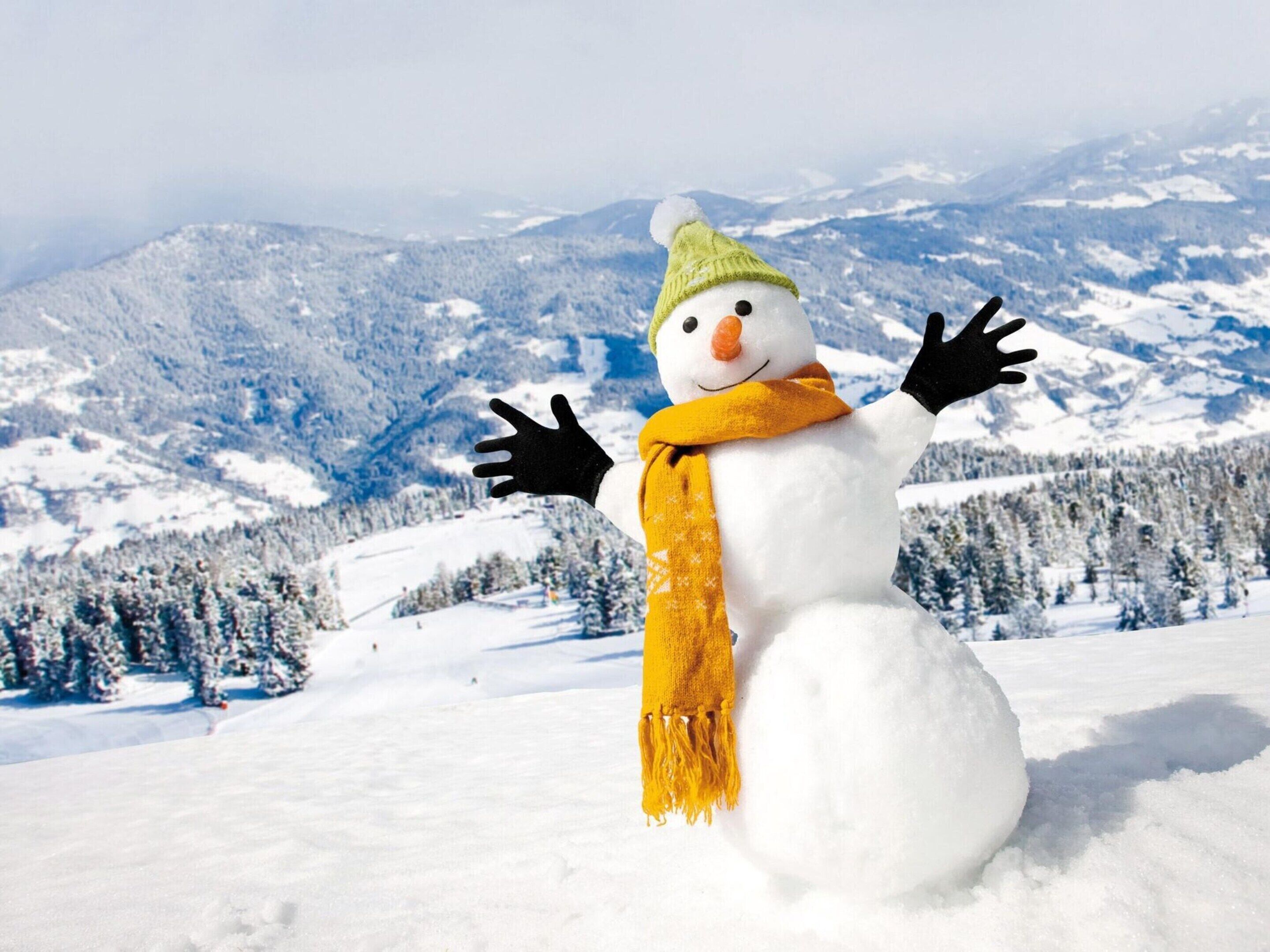 A snowman with a green hat and yellow scarf in a snowy landscape with mountains in the background.