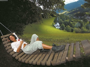 A person lying on a wooden swing chair overlooking a rural landscape with a church and houses. - Cozy Apartment near Ski Area in Bad Kleinkirchheim (Bad Kleinkirchheim)