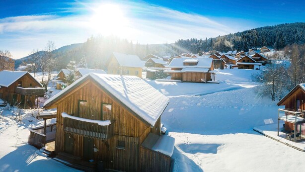 Eine Gruppe Holzhütten mit schneebedeckten Dächern, eingebettet in eine verschneite Landschaft unter blauem Himmel.