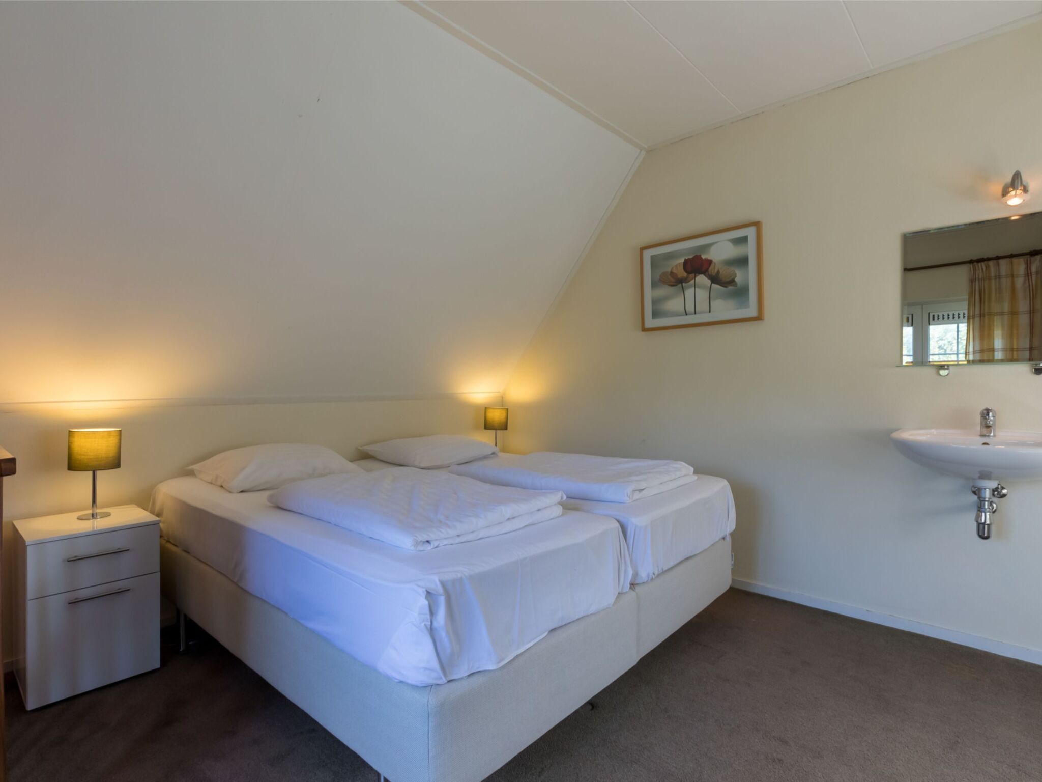 A bedroom with a bed, bedside tables, a lamp, a painting, a mirror, and a sink.