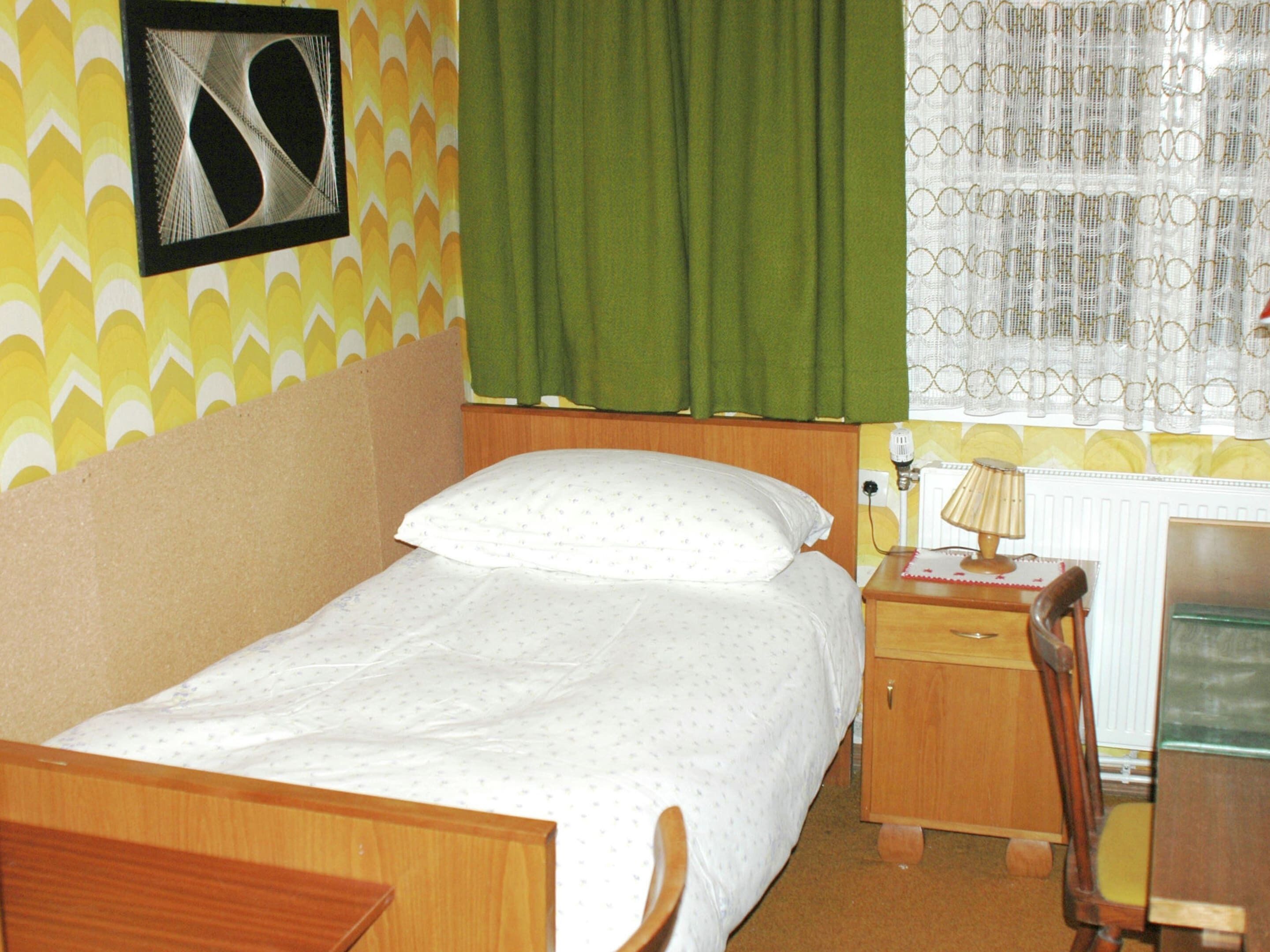 A small, single-bed room with a wooden headboard, a bedside table with a lamp, and a chair.