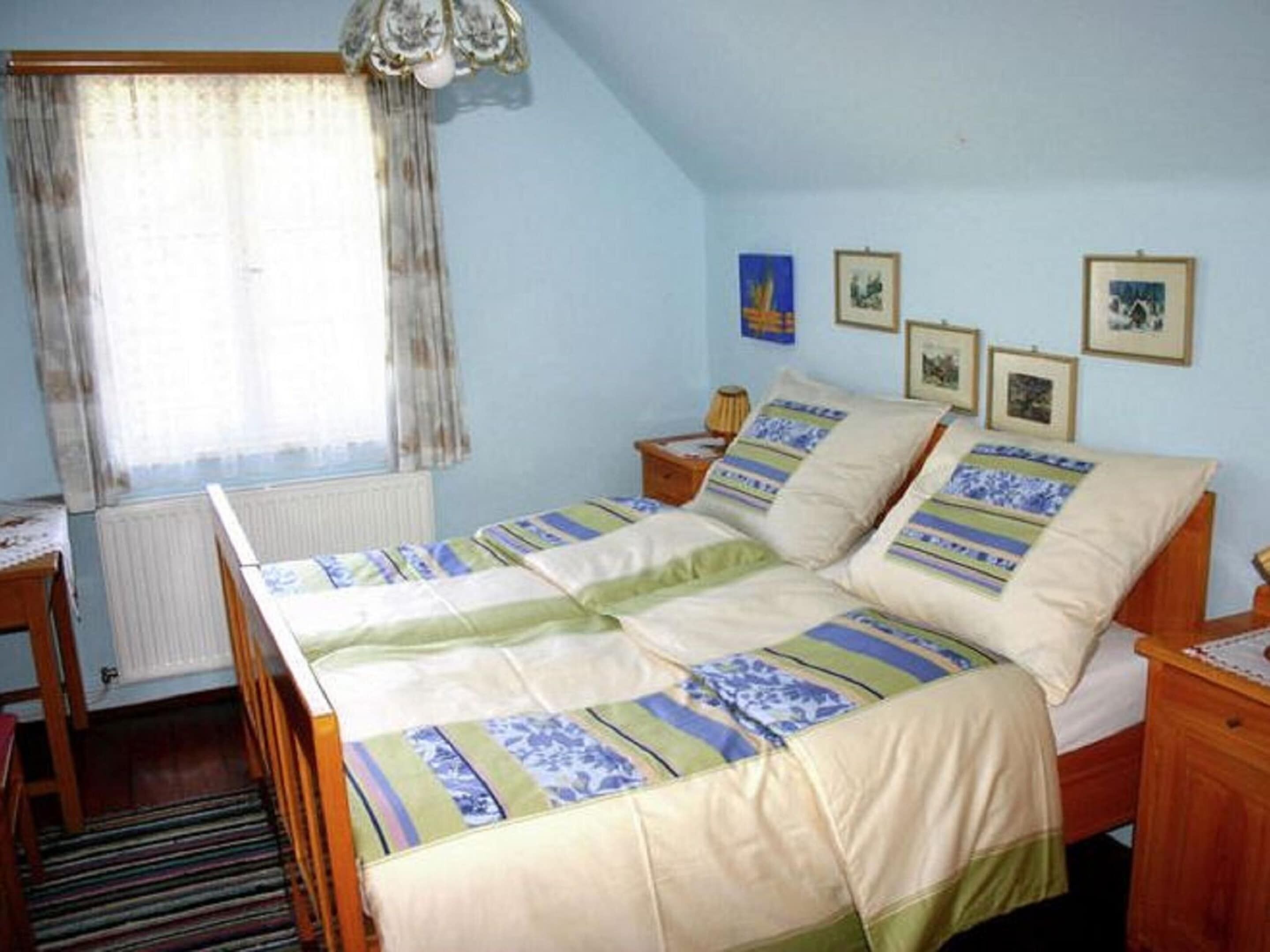 Une chambre à coucher avec un lit, une fenêtre avec des rideaux, un radiateur, une table de chevet en bois et des tableaux encadrés accrochés au mur.