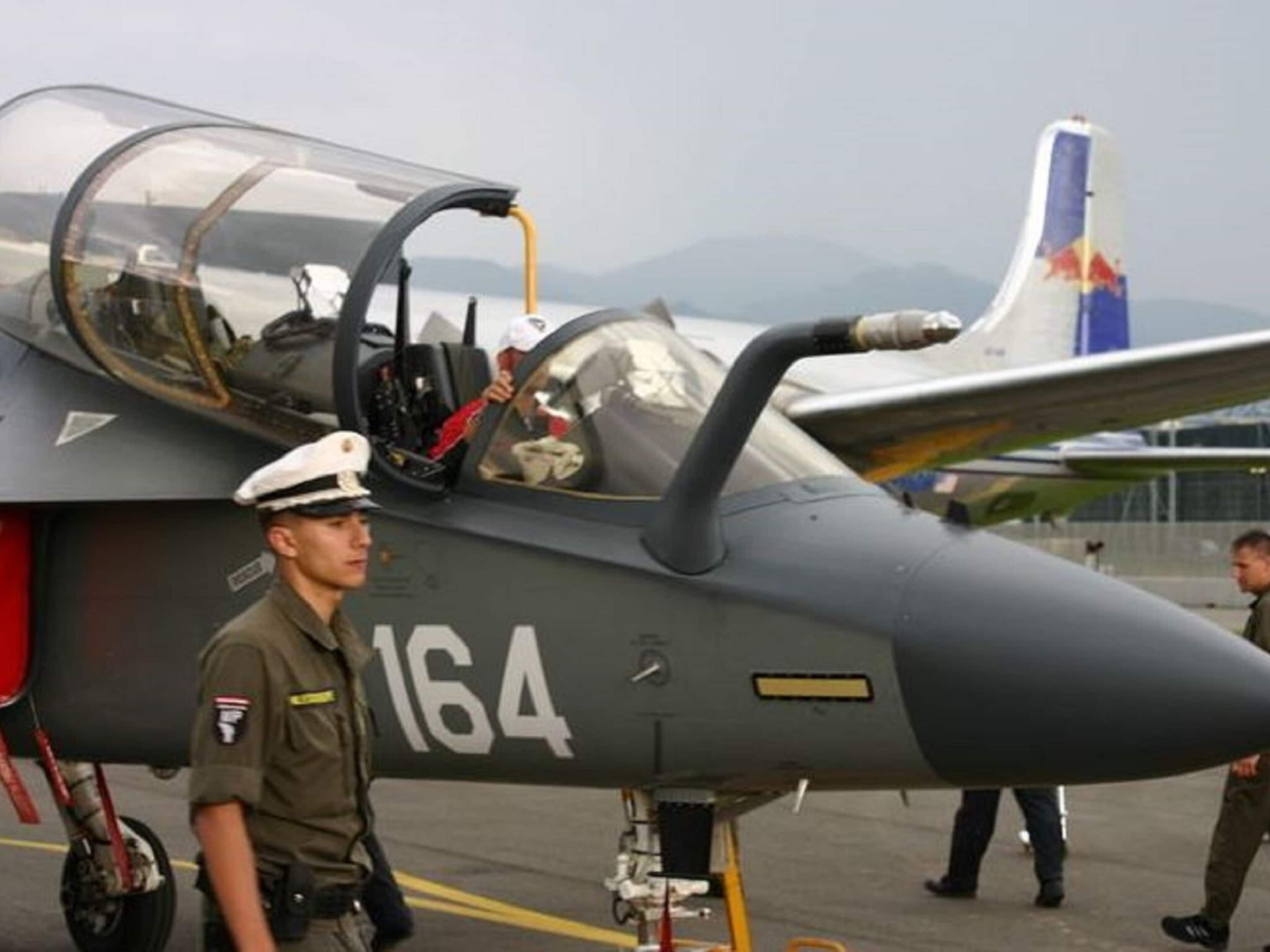 Un avión de combate con el número 164 en la parte lateral, estacionado sobre una pista con montañas al fondo.