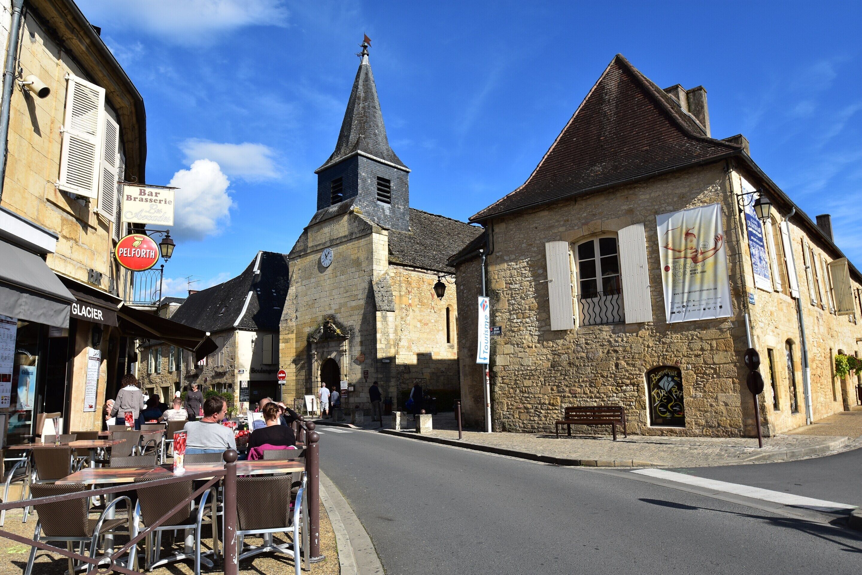 Une rue bordée d’immeubles en pierre, avec un clocher d’église, des tables en terrasse et un café appelé « Bar Brasserie ».