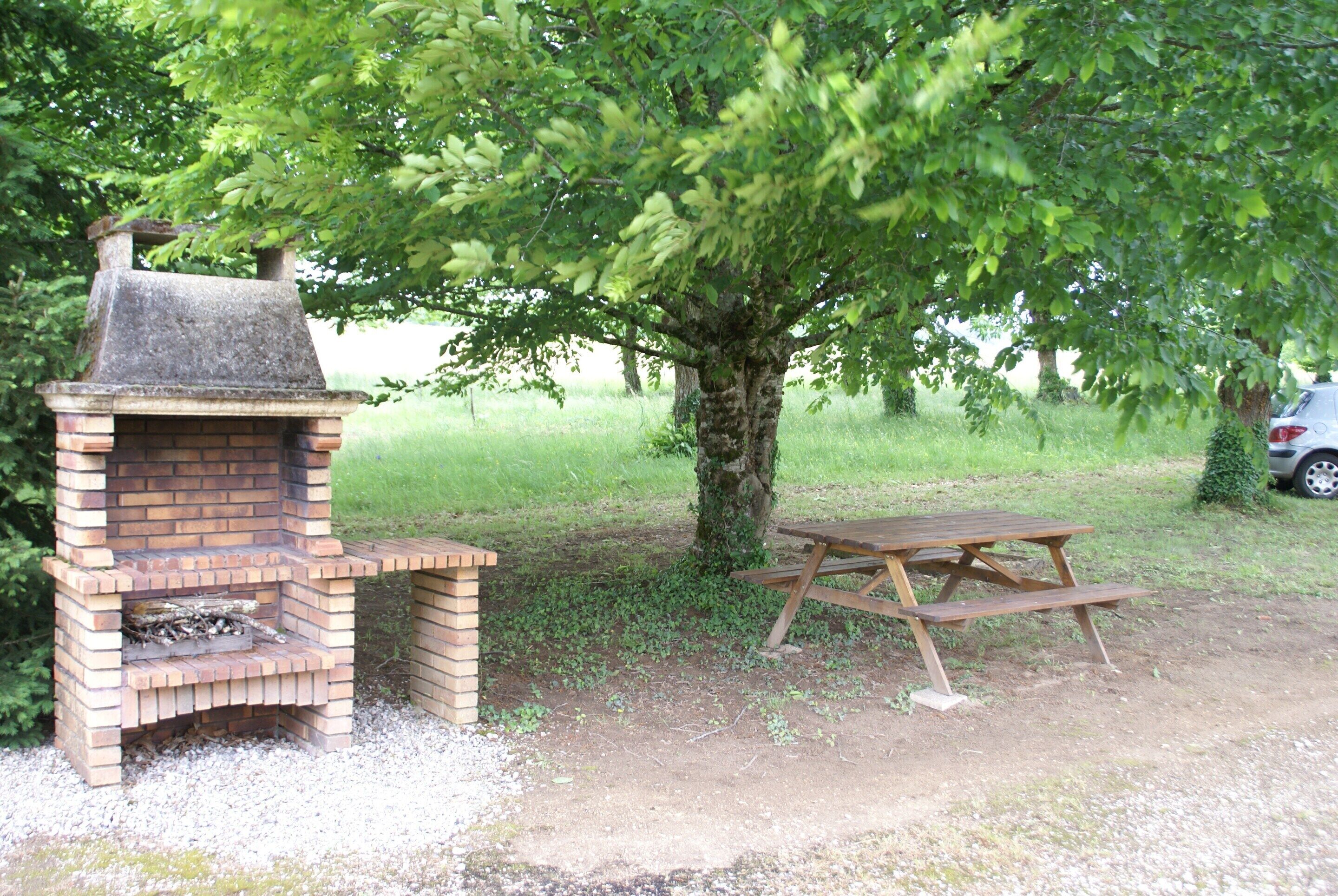 En udendørs grill i mursten og et picnicbord under et træ.