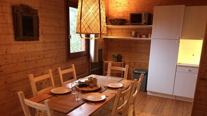 A wooden cabin interior with a dining table set for four, wooden chairs, a microwave, a refrigerator, and a window with a view of trees. - Lakeside Escape for 6 Guests (Beaulieu)