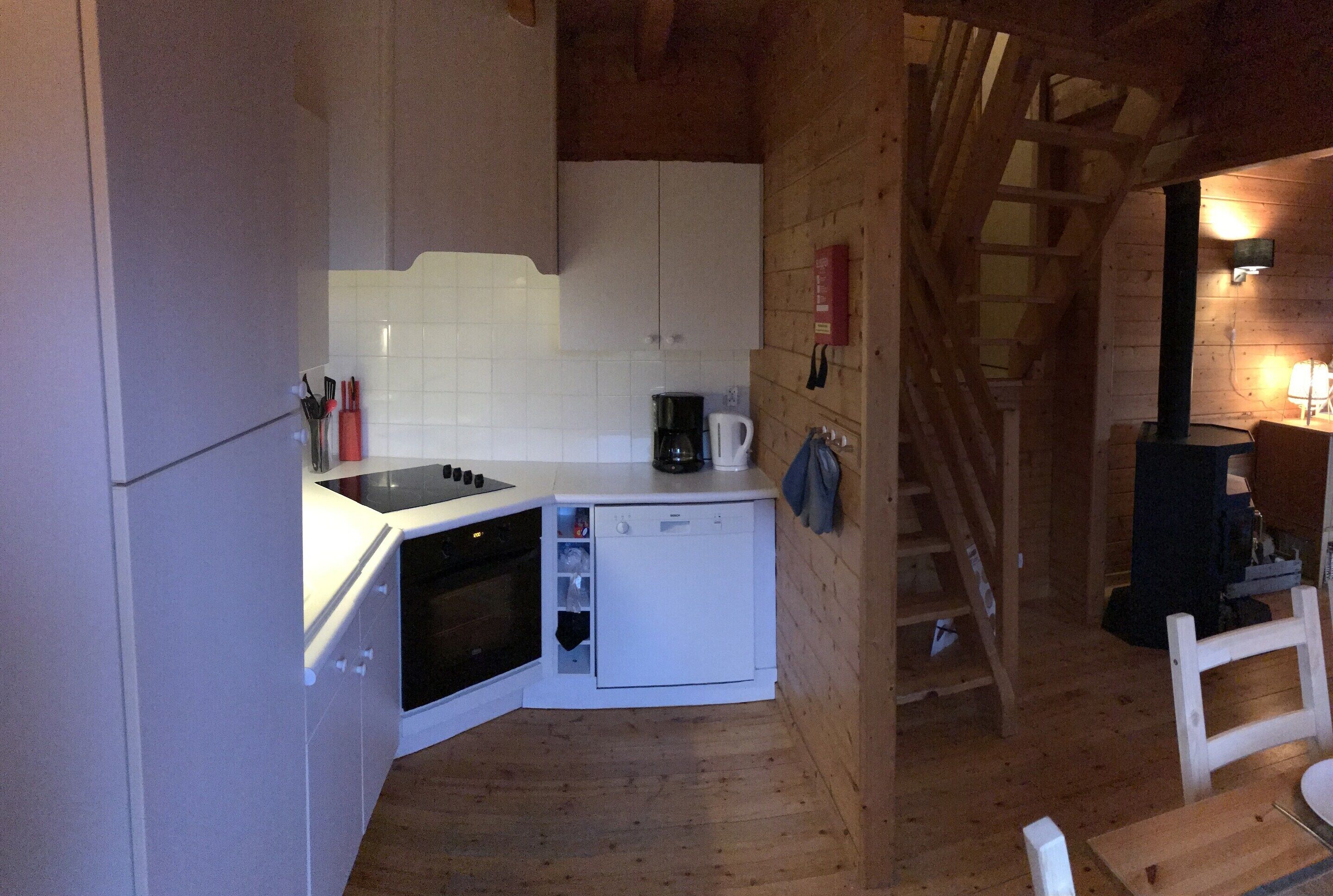 Une cuisine compacte avec des meubles blancs, un lave-vaisselle et une cuisinière. Un escalier en bois mène à une autre pièce.