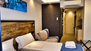 Habitación de hotel con cabecera de madera, cama con sábanas blancas, manta azul, almohada a rayas, aire acondicionado de pared y una pequeña área de kitchenette.