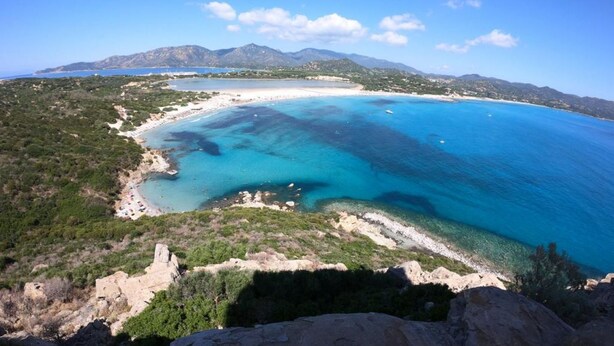 Eine Küstenlandschaft mit Strand, klarem blauen Wasser und Bergen im Hintergrund.