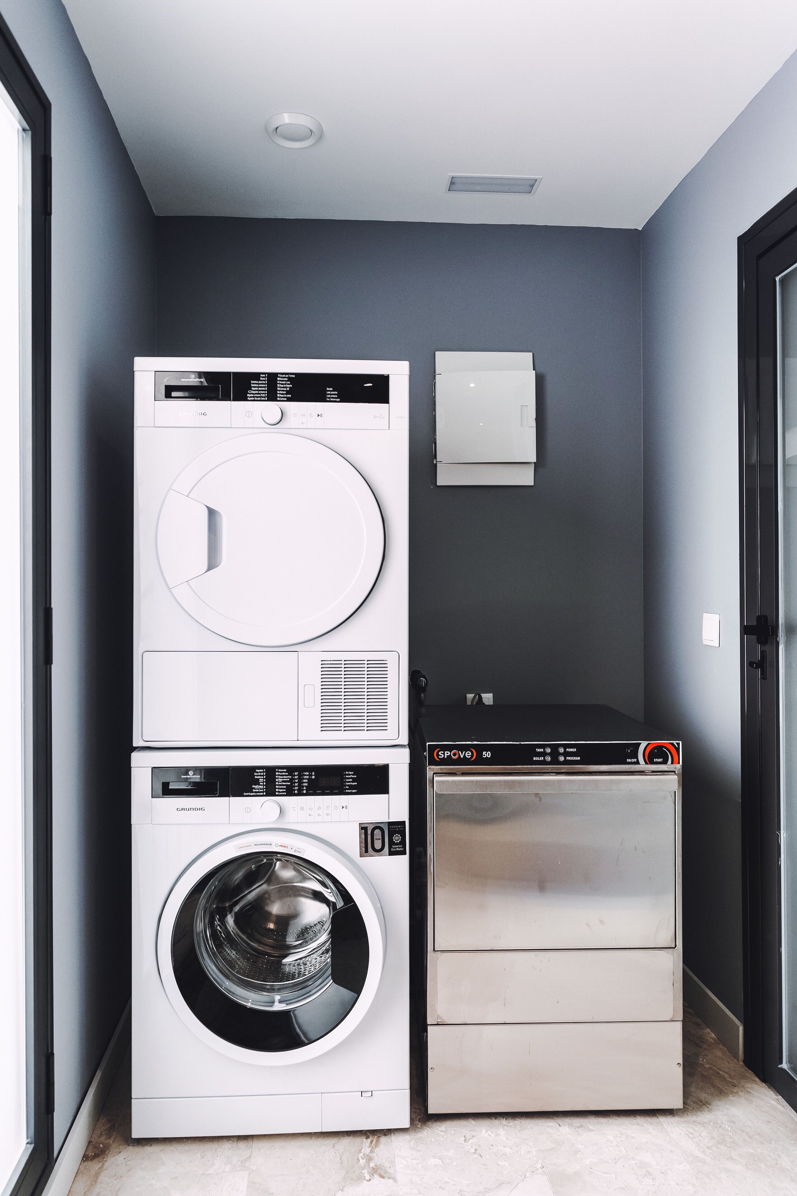 A compact laundry room with a stacked washer and dryer, a small wall-mounted heater, and a door to the right.