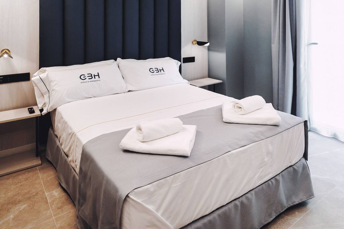 Un cuarto de hotel con una cama bien tendida, una mesita de noche con l ámpara y vista al exterior por la ventana.