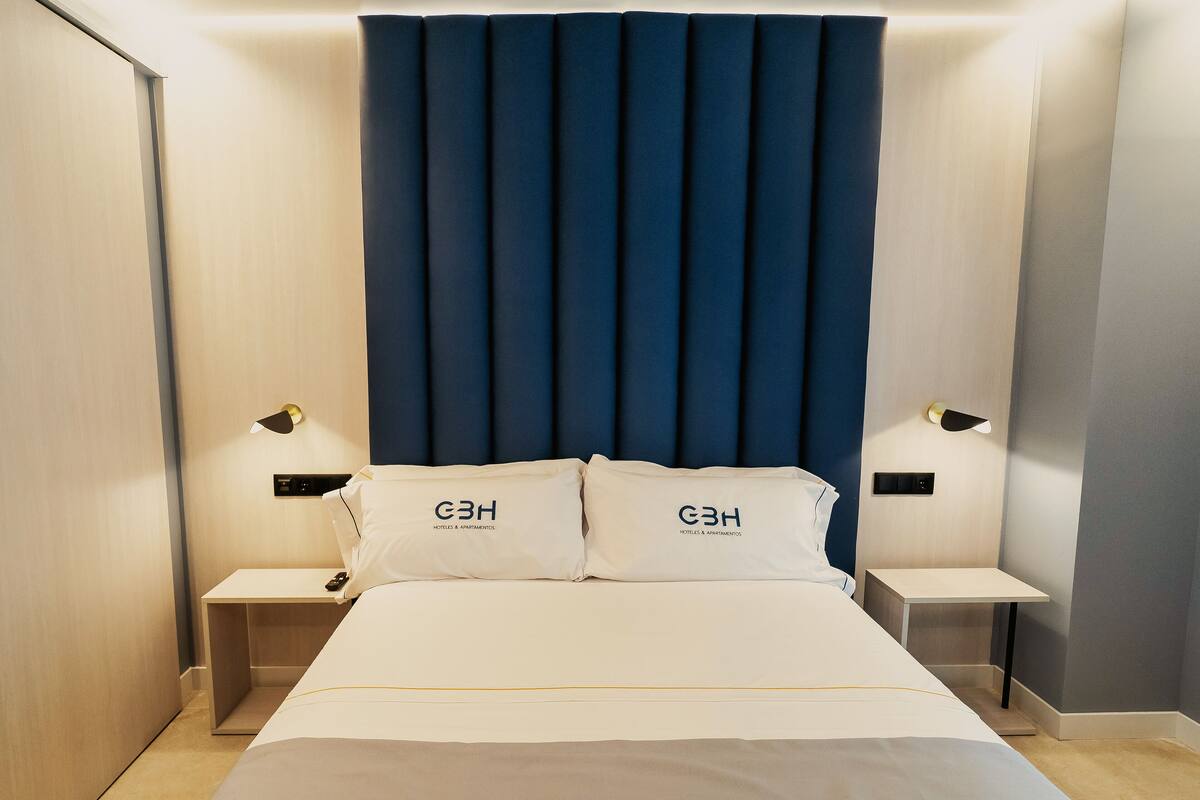 Una habitación de hotel con una cama grande, dos mesitas de noche y un cabecero con líneas verticales.