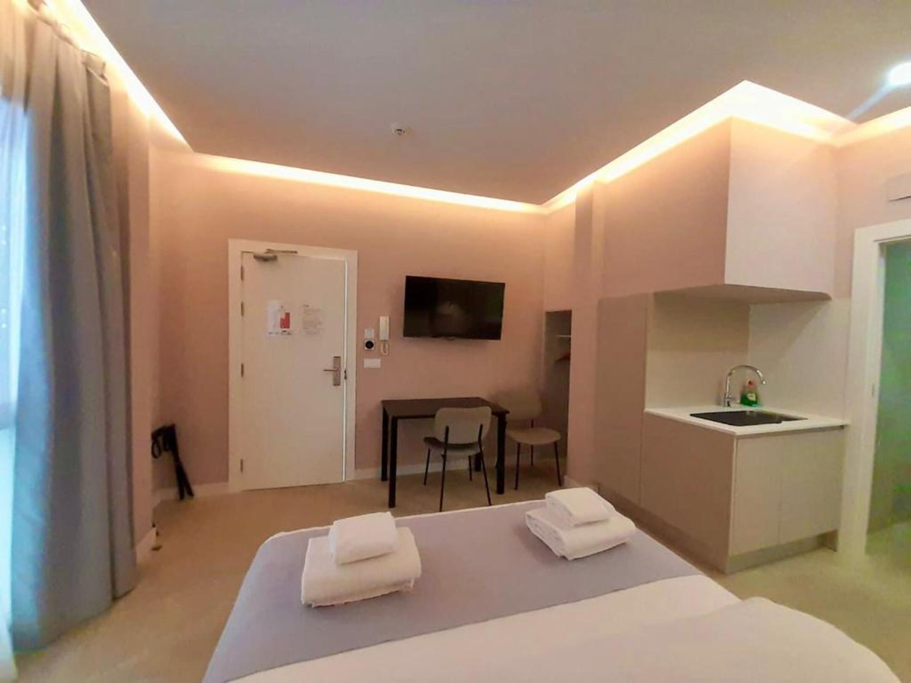 A hotel room with a bed, a small table, a chair, a TV, and a kitchenette.