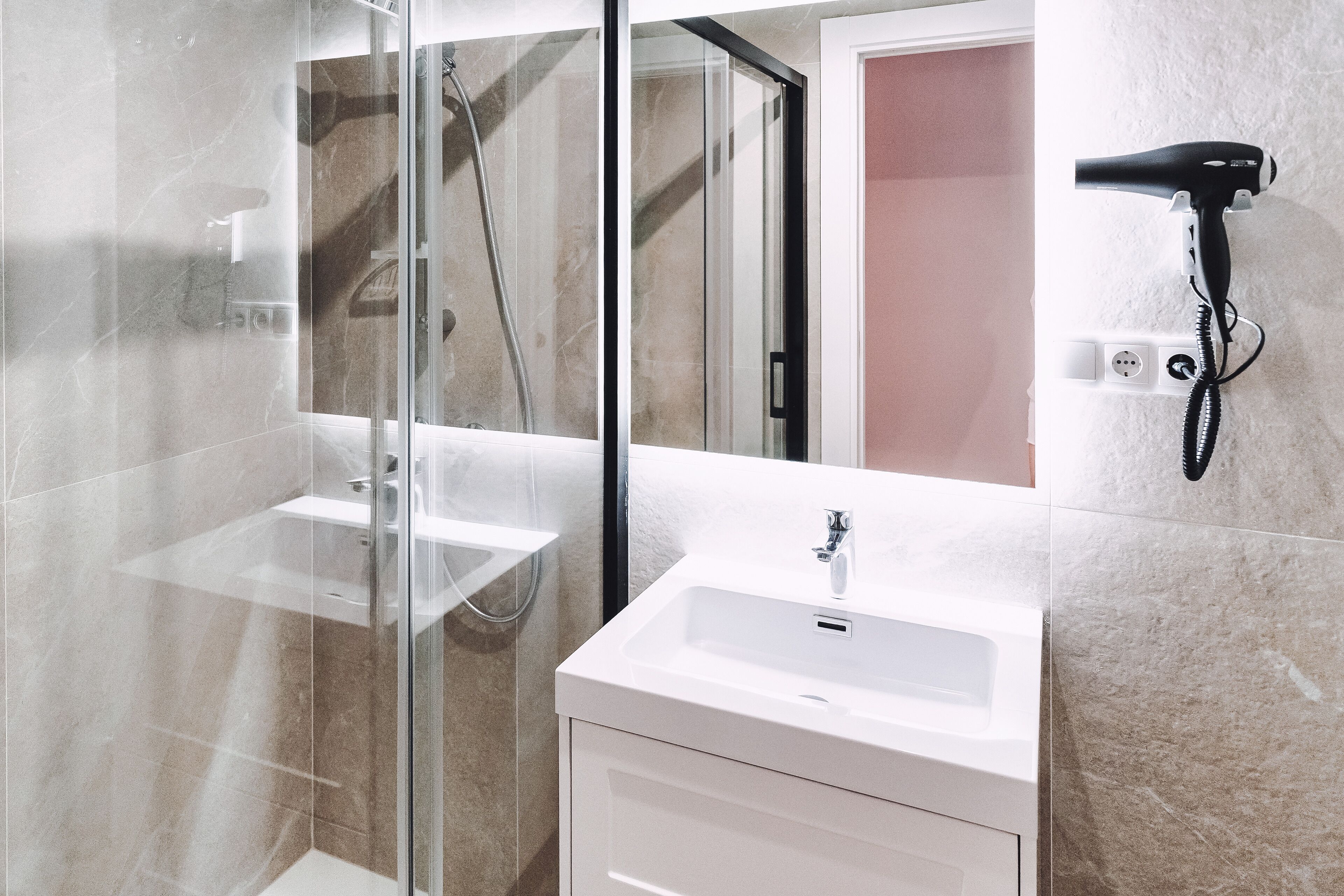 A modern bathroom with a glass shower enclosure, a white sink, and a hairdryer mounted on the wall.