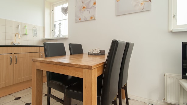 A dining area with a wooden table and black chairs, a kitchen with light-colored cabinets, and a window with a view. - Apartment in a Quiet, Rural Area, not far From the Centre (Callantsoog)