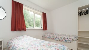 A bedroom with two beds, a window with red curtains, a radiator, and a shoe rack. - Apartment in a Quiet, Rural Area, not far From the Centre (Callantsoog)