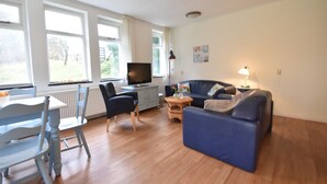 A living room with a blue sofa, a television, a dining table with chairs, and large windows. - Timeless Charm and Cozy Stay (Bergen aan Zee)