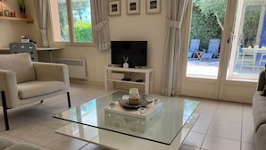 Ein Wohnzimmer mit Glastisch, zwei Sofas, einem Fernseher und einem Essbereich, der durch eine Glastür sichtbar ist.
