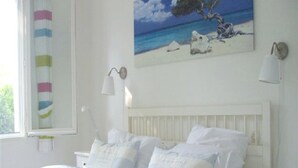 Ein Bett mit weißer Bettwäsche und einer grünen Tagesdecke, ein Strandbild und ein Fenster mit einem gestreiften Vorhang.