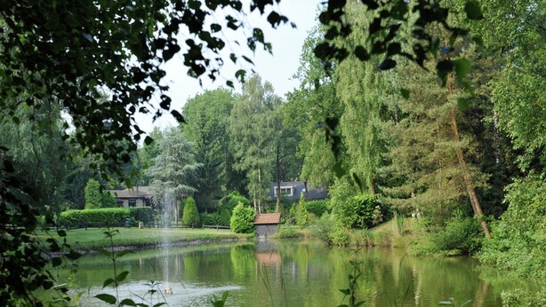 A serene pond with a fountain, surrounded by lush greenery and a house in the background. - Chalet in Holiday Park With Private Terrace (Baarle-Nassau)