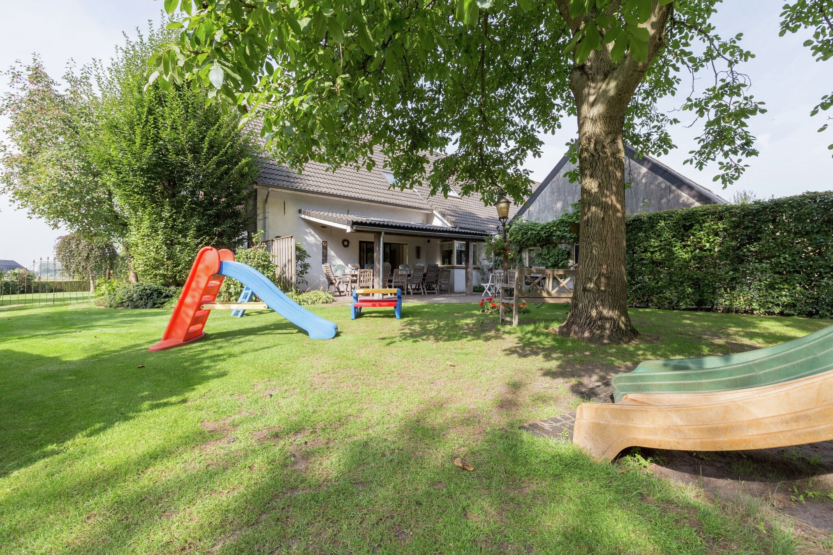 Ein grünes Hinterland mit Spielbereich, darunter eine Rutsche und eine Schaukel, ein Baum und ein Haus mit Veranda.