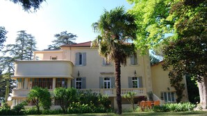 A multi-story building with a curved facade, balconies, and a well-maintained garden with trees and flowers. - Mansion in Provence With Pool & Near Town (Valreas)