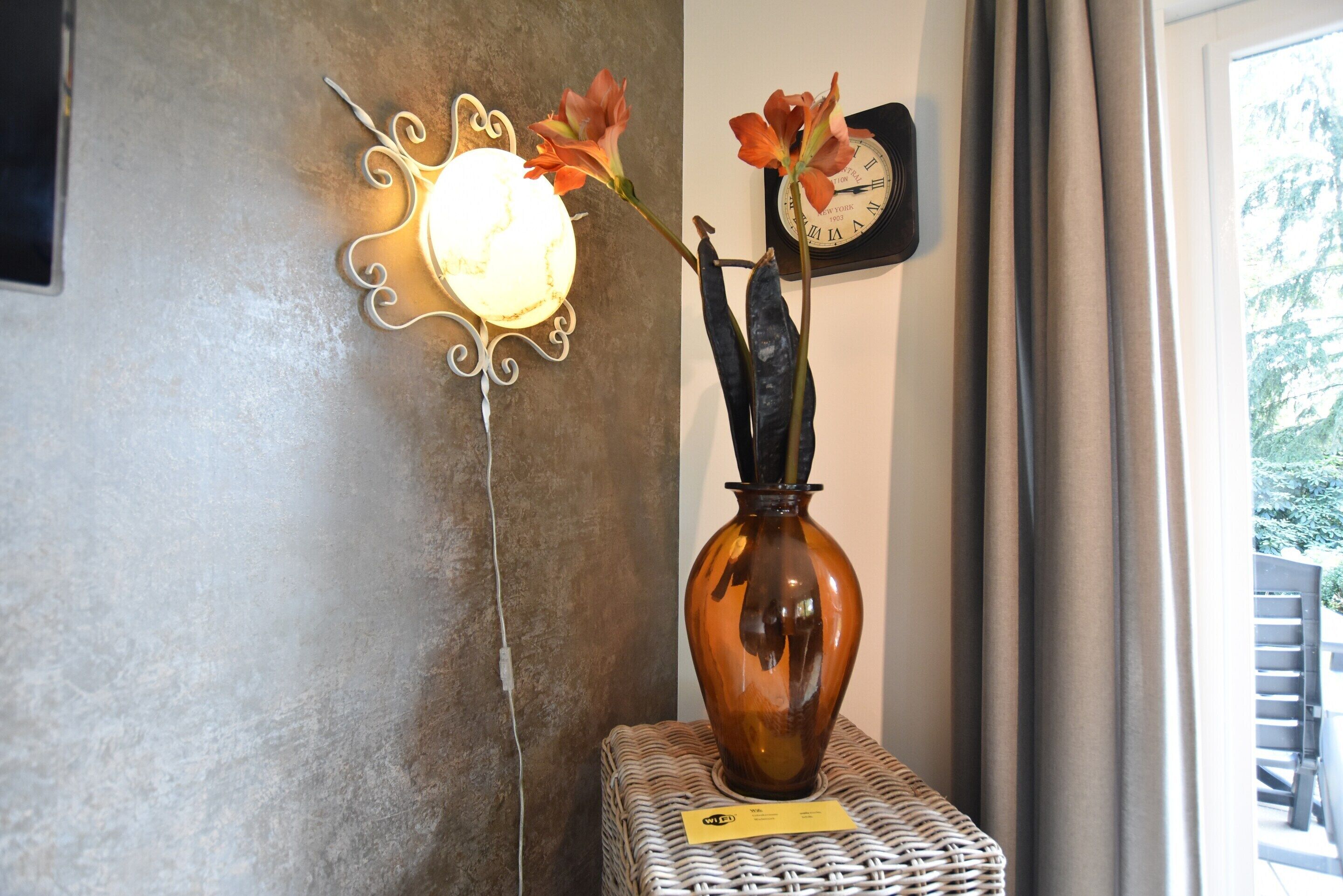 Eine Vase mit Blumen auf einem Tisch, eine Wanduhr und eine Zierleuchte.
