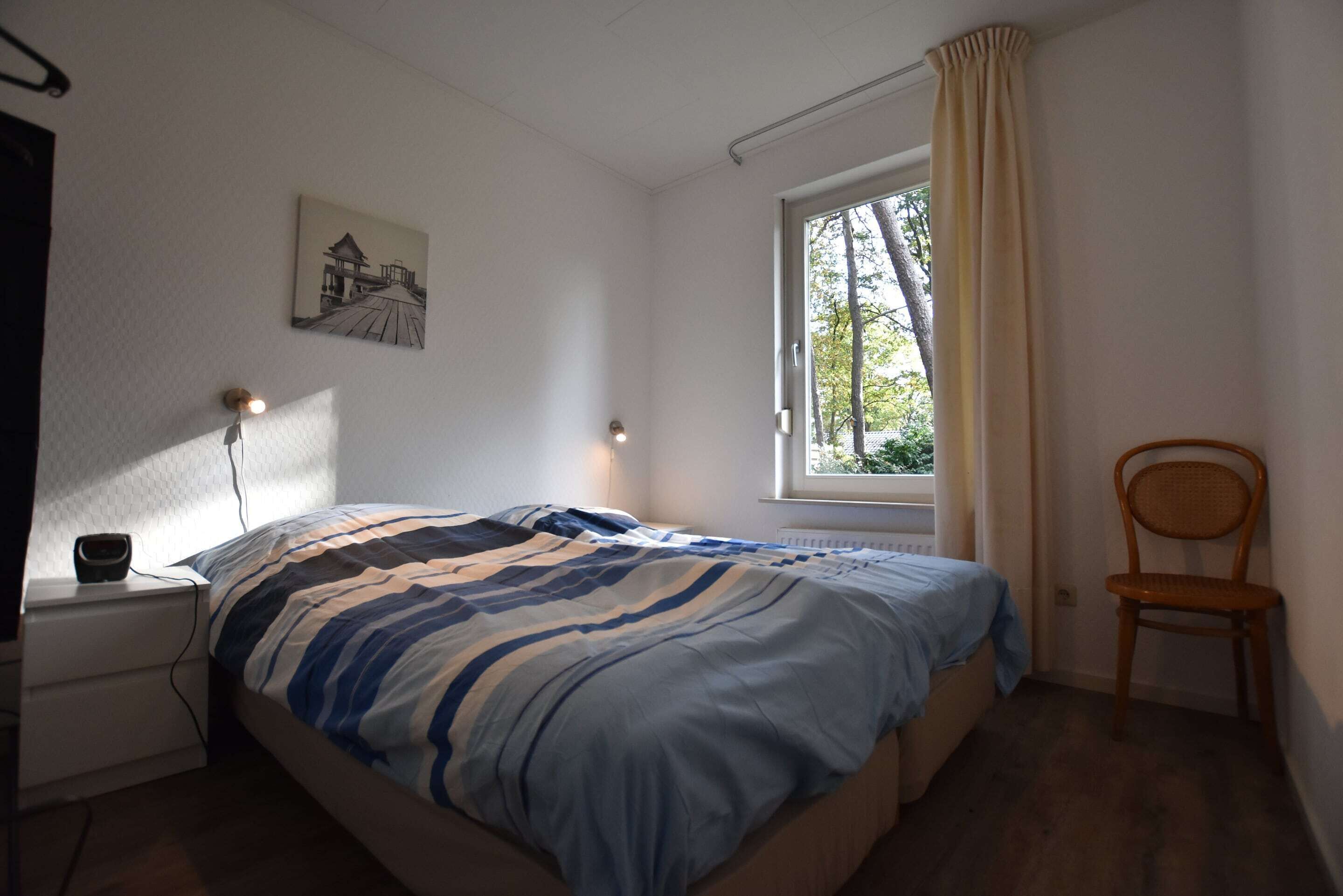 A bedroom with a bed, bedside table, lamp, chair, window, and a picture on the wall.