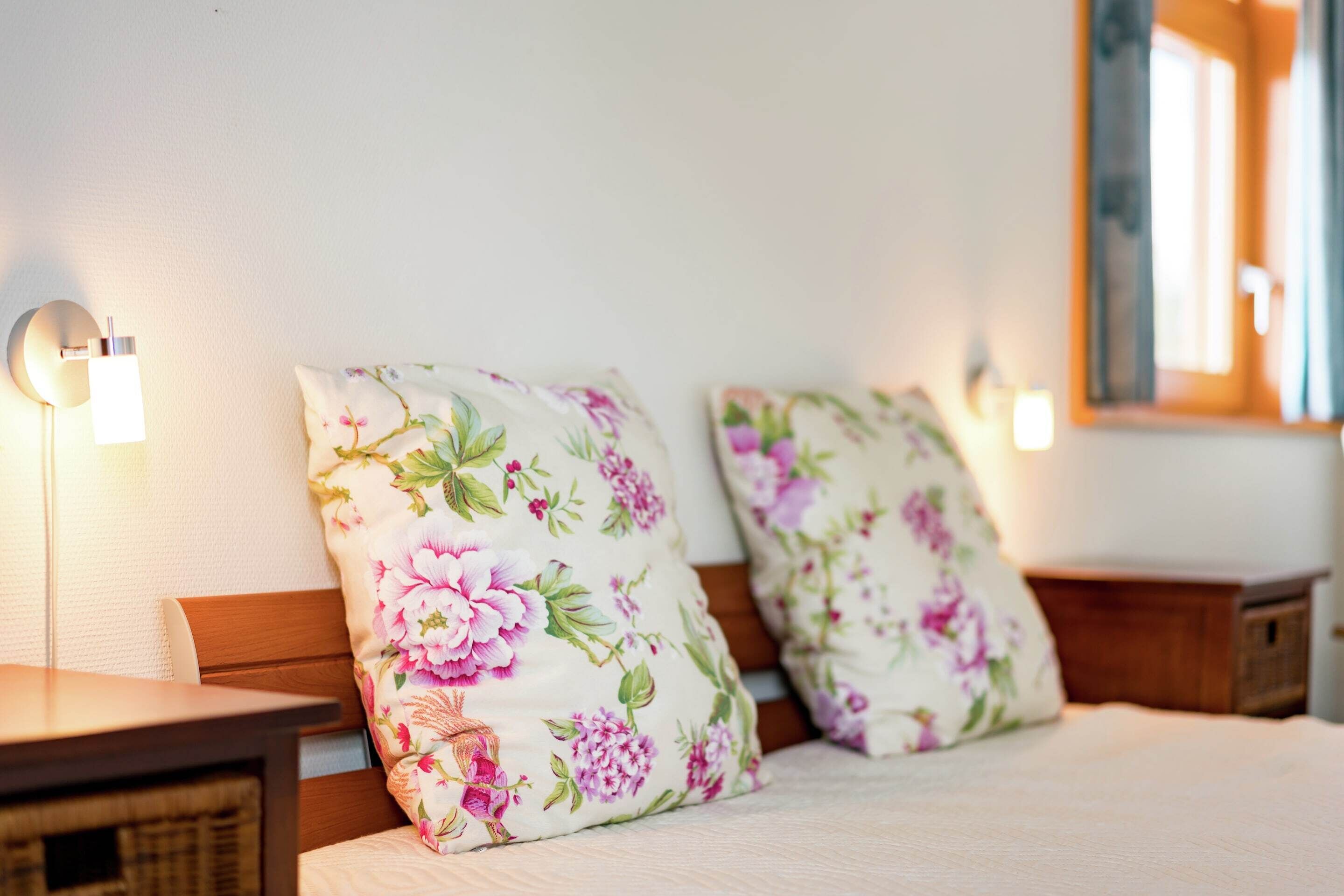 Ein Schlafzimmer mit zwei Nachttischen aus Holz, einem Bett mit blumigem Kopfkissen und einem an der Wand befestigten Spiegel.