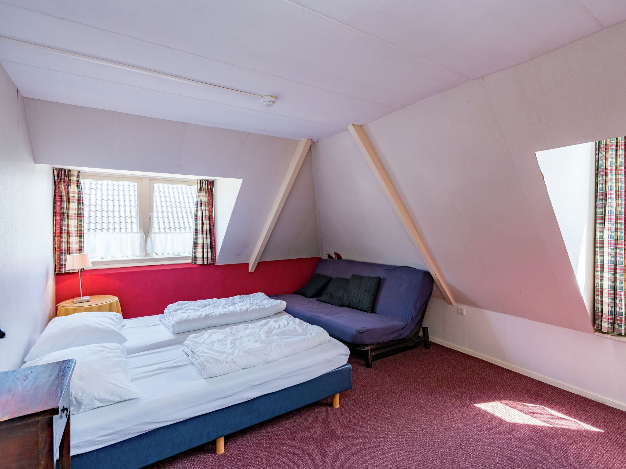 Ein Schlafzimmer mit Bett, einem Fenster mit Vorhängen, einer roten Wand und einer geneigten Decke.