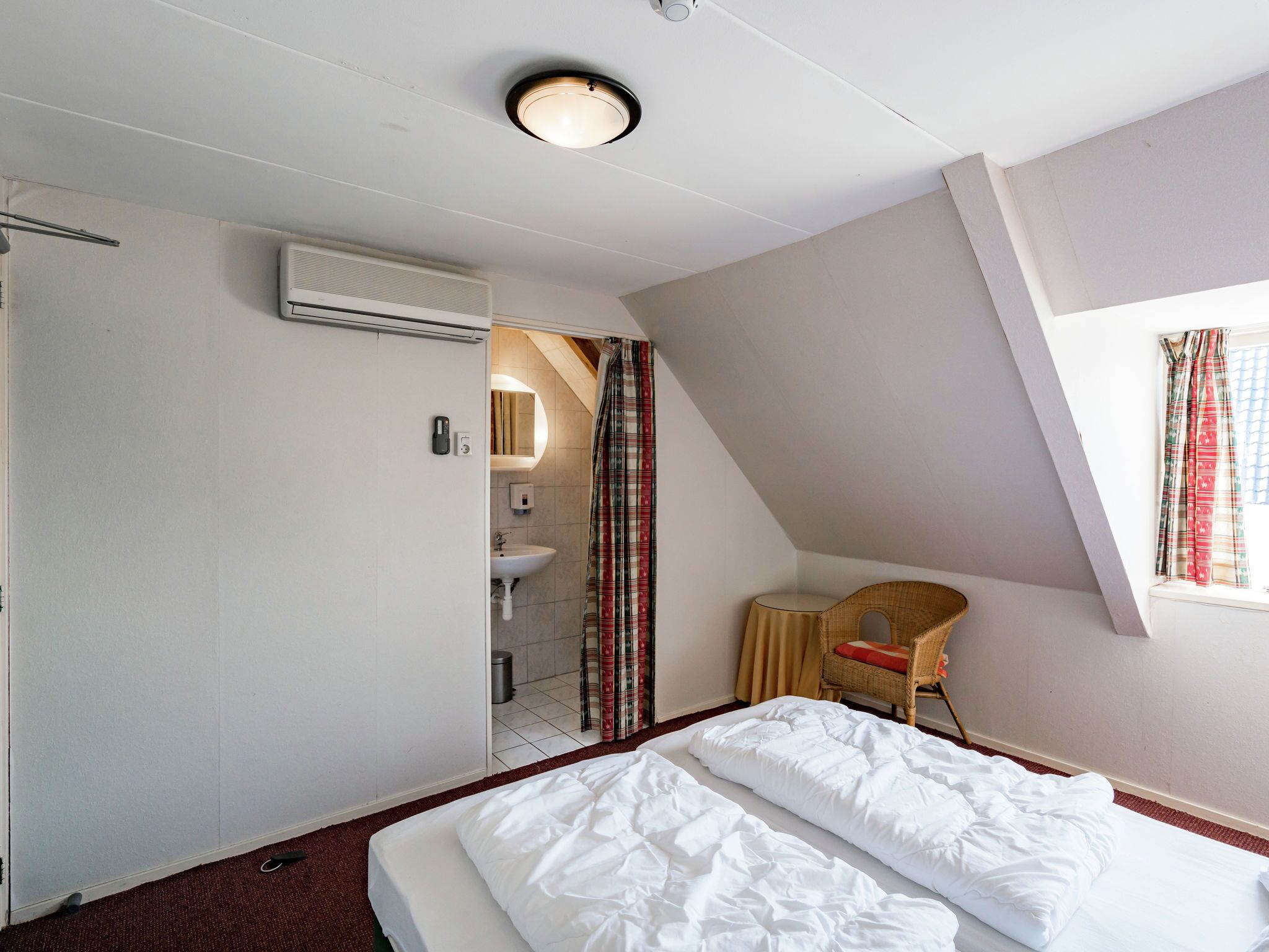 A hotel room with a bed, a small desk, a chair, a window with curtains, a bathroom with a sink and mirror, and an air conditioning unit.