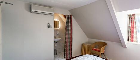 A hotel room with a bed, a small desk, a chair, a window with curtains, a bathroom with a sink and mirror, and an air conditioning unit.