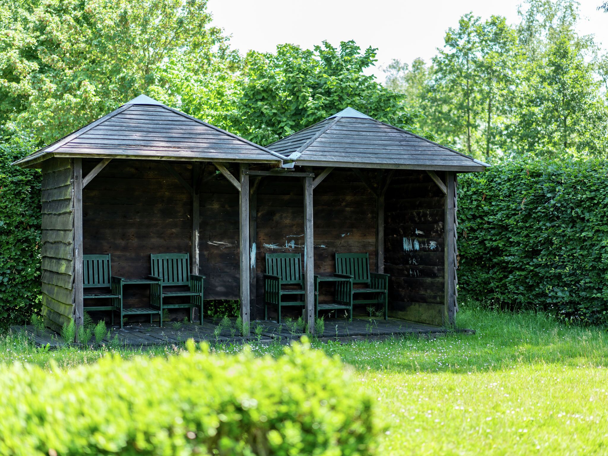 A rustic wooden structure with two covered seating areas, each with a bench and a chair.