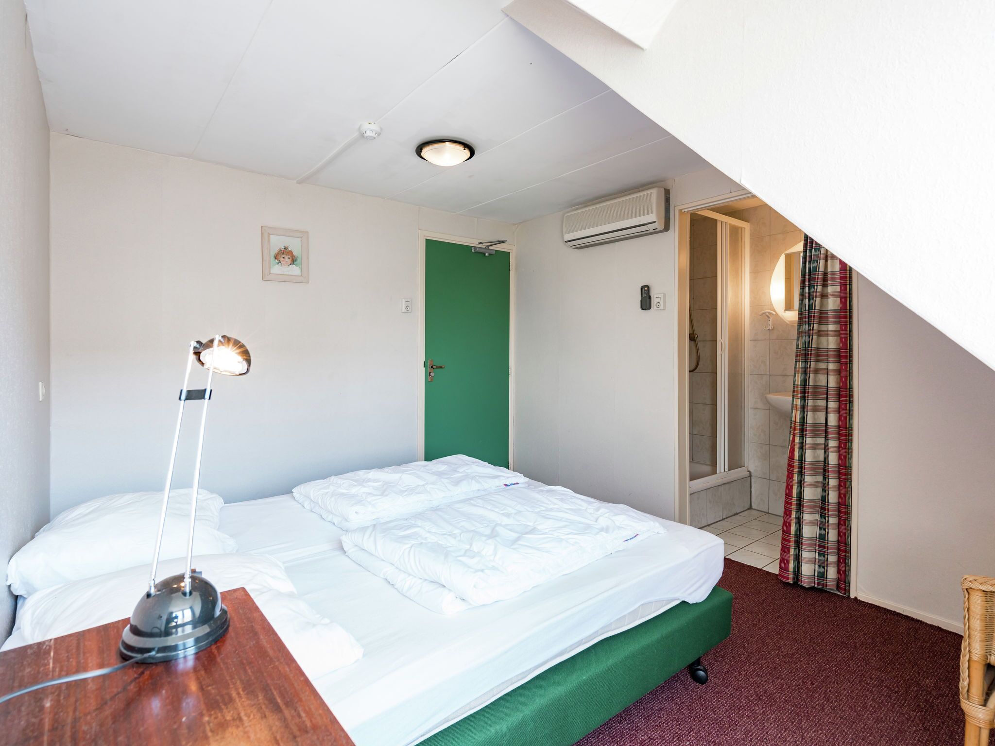 A bedroom with a bed, a desk with a lamp, a green door, an air conditioning unit, and a shower area.