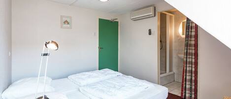 A bedroom with a bed, a desk with a lamp, a green door, an air conditioning unit, and a shower area.
