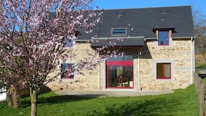 A stone building with a red-framed door and windows, a sloped roof, and a tree in the foreground. - Family Escape With Terrace (Isigny-le-Buat)