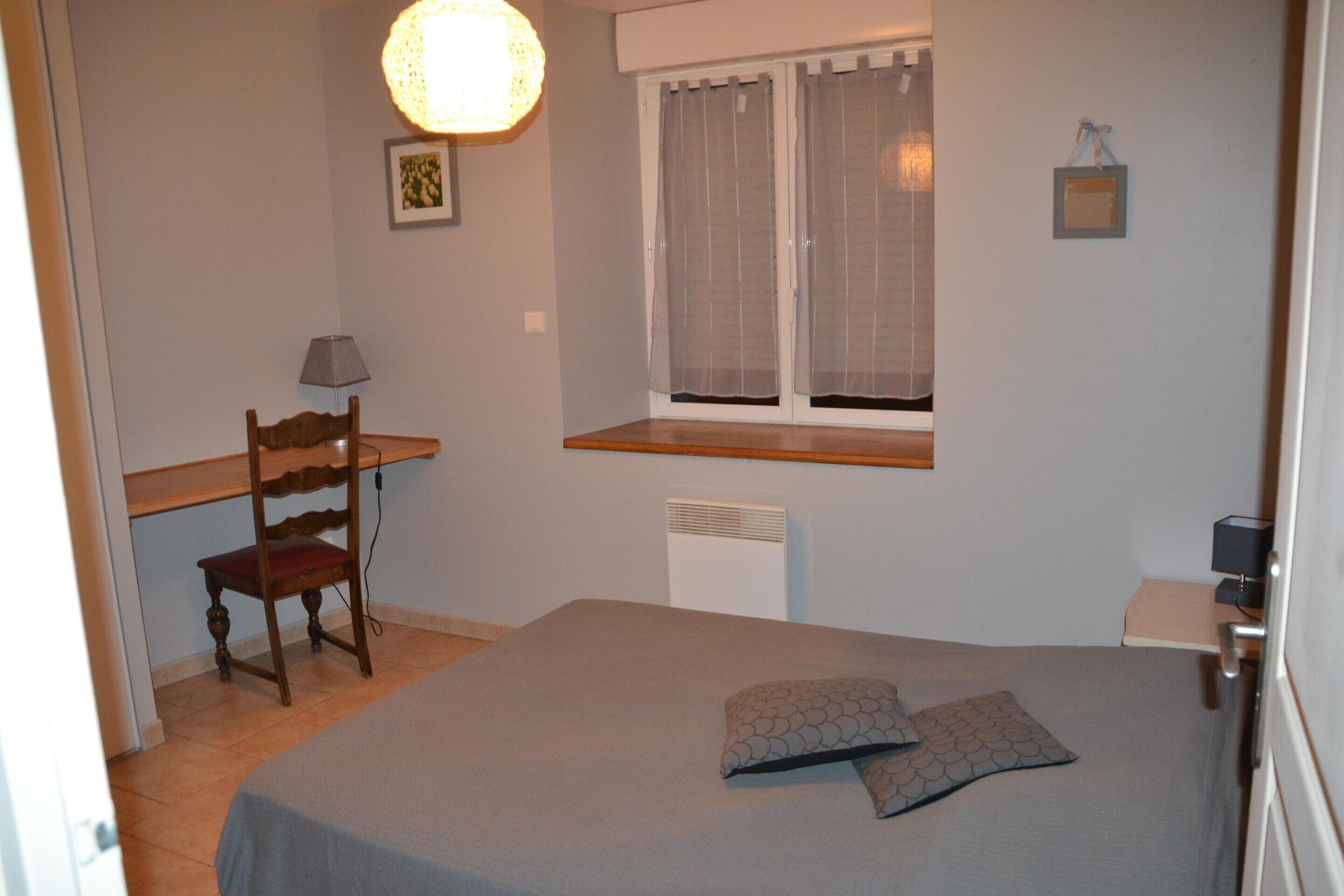 A bedroom with a bed, a chair, a desk, a window with curtains, and a wall-mounted lamp.