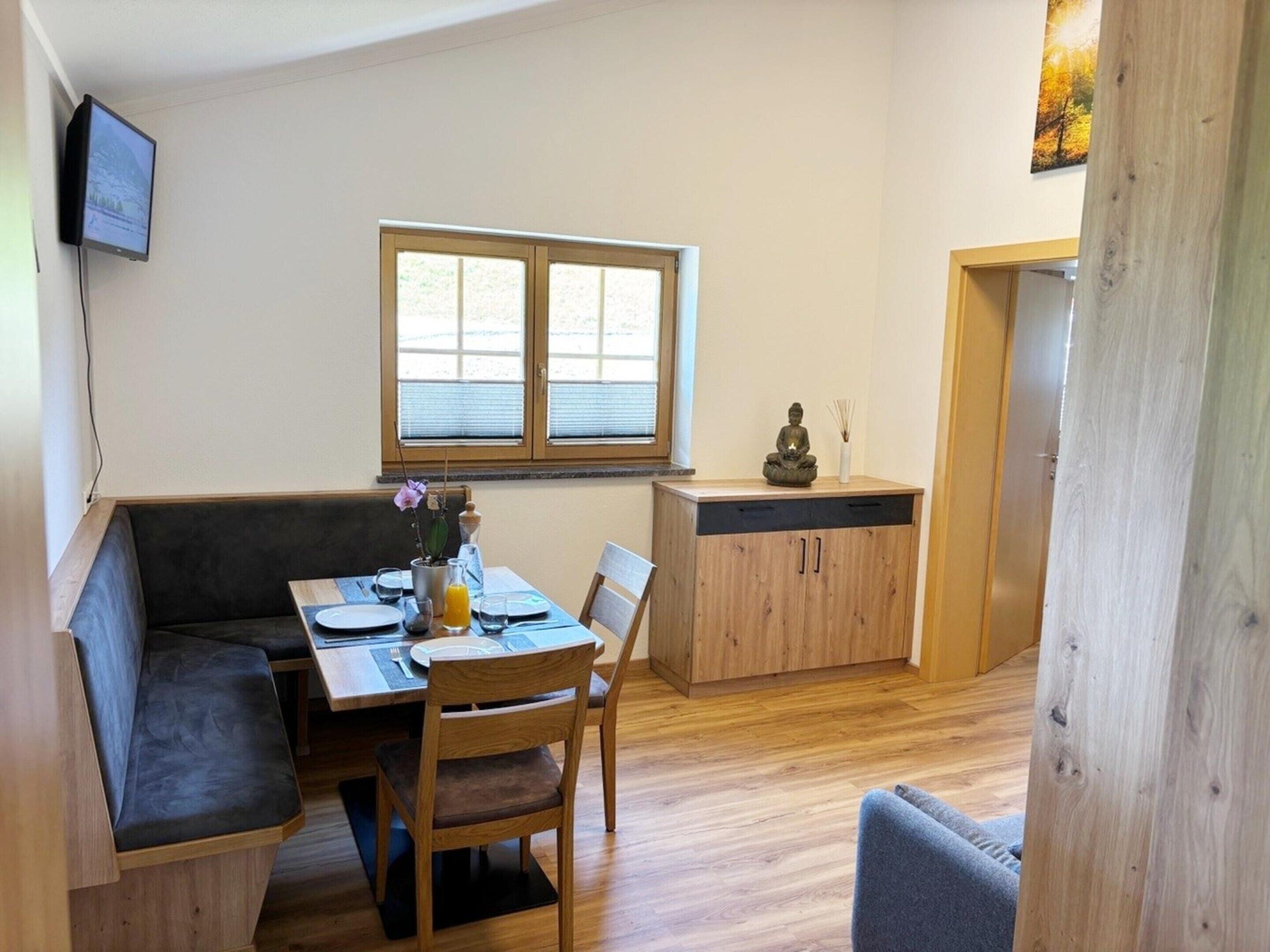 A cozy dining area with a wooden table, cushioned bench, and chairs. A window with blinds is above the table, and a small statue is on a cabinet.