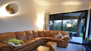 Ein modernes Wohnzimmer mit einem braunen Ledersofa, zwei Holztischen und einem großen Fenster mit Blick auf Grünflächen.